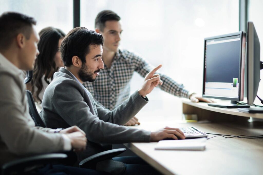 A photo of a group of people looking at a computer screen, one person is pointing to the screen with one hand, with the other hand on the mouse of a computer.